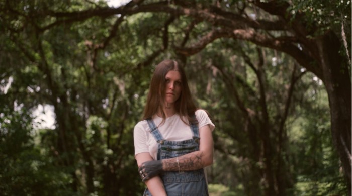A person with long brown hair wearing a white tshirt and blue denim overalls in a woods